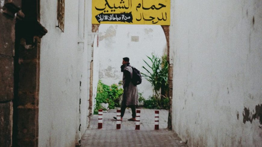 Man walking through a rustic alleyway with Arabic signage, surrounded by white walls and vegetation.